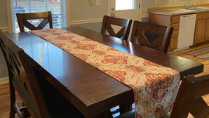 A dining area with a long wooden table, chairs, a kitchen in the background, and a 'Gather Together & Give Thanks' sign on the wall. - Quiet, Cozy, and Family Friendly in Hendo (Hendersonville)