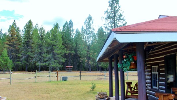 A rustic cabin with a porch, surrounded by a fenced-in grassy area and dense forest. - "The Old Homestead"  An Original Homestead Cabin (Trout Creek)