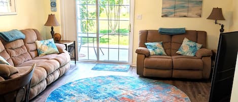 A living room with two sofas, a coffee table, a TV stand, a ceiling fan, and a large window with a view of the outdoors.
