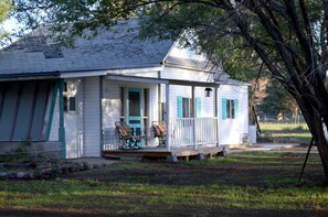 A white house with blue shutters, a porch with two chairs, and a tree in the background.
