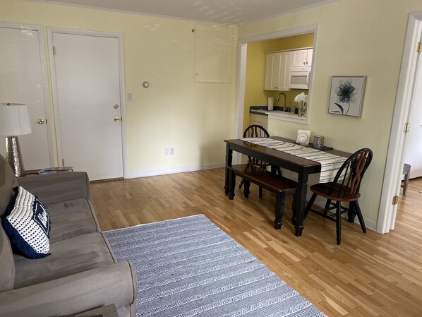 A living room with a sofa, a patterned rug, a dining table with chairs, and a kitchen area with cabinets and a microwave.