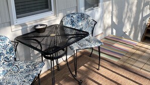 A covered porch with a table and chairs, a window with blinds, and a door with a number plate.