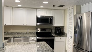 A modern kitchen with white cabinets, stainless steel appliances, granite countertops, and two black bar stools. - Beautiful Beach! Spectacular Sunsets! (Redington Shores)