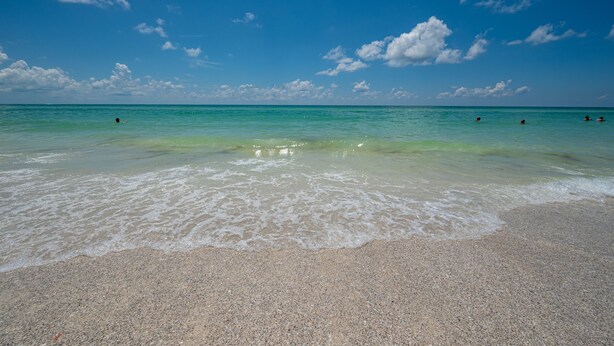 A beach with clear turquoise water, gentle waves, and a few people swimming.
