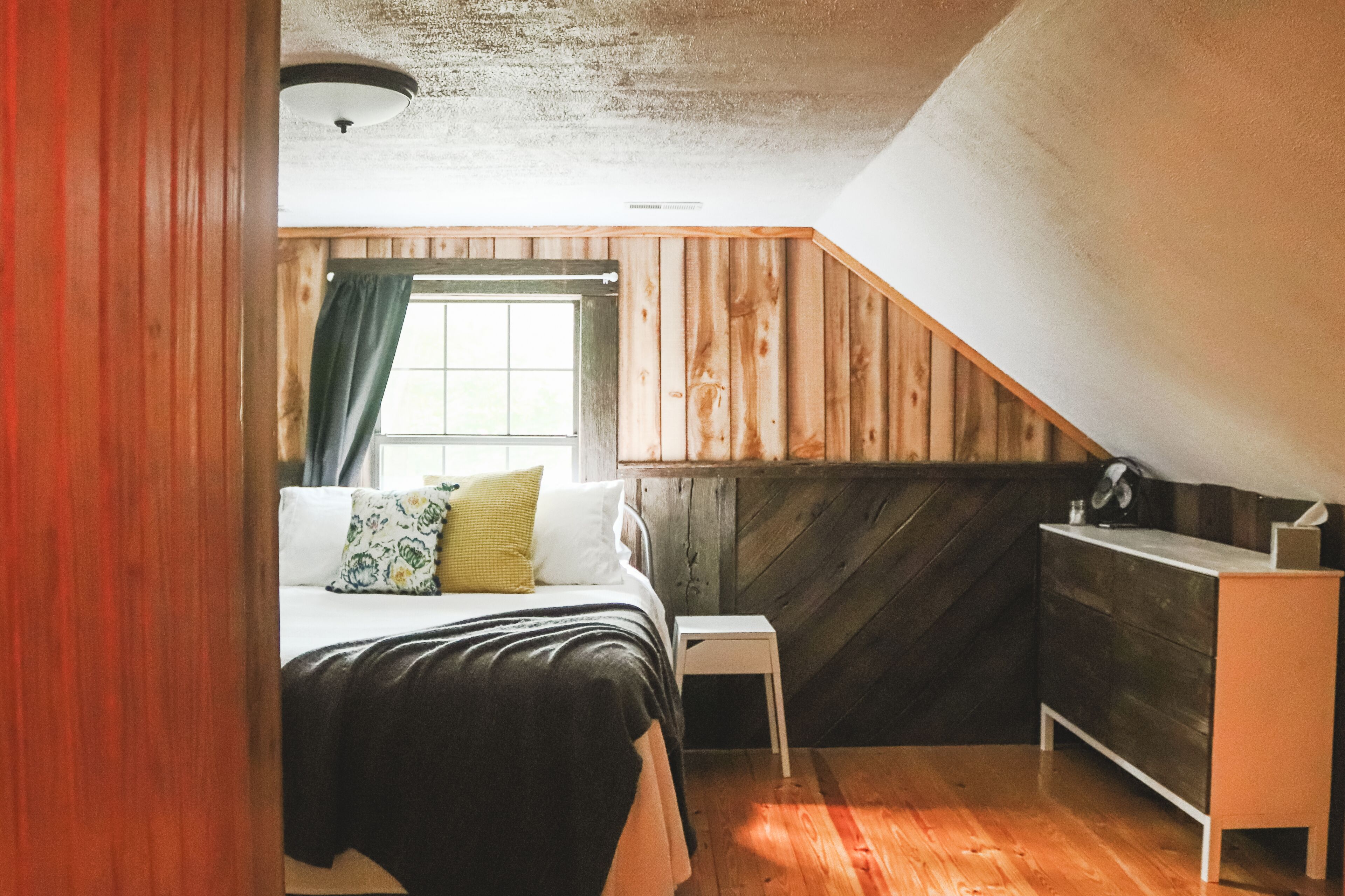 Un dormitorio con cama, una mesita de noche pequeña, un aparador y una ventana con cortinas.