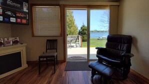 A room with a wooden floor, a black leather chair, a wooden chair, a fireplace, a TV mounted on the wall, and a sliding glass door leading to a deck with a view of a lake.