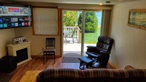 A living room with a fireplace, a television mounted on the wall, a sofa, and a chair. There is a sliding glass door leading to a balcony with a view of greenery and a body of water.
