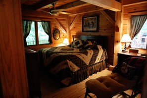 A cozy log cabin bedroom with a bed, a chair, a fireplace, and a window with curtains. - Lake Superior Beachfront, 4+ Bedrooms, Spectacular Timberframe Home (Washburn)