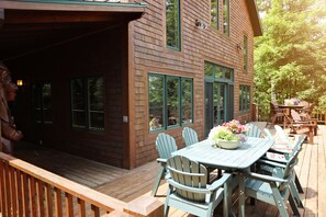 A wooden deck with a table and chairs, a covered area, and a view of a forested area. - Lake Superior Beachfront, 4+ Bedrooms, Spectacular Timberframe Home (Washburn)