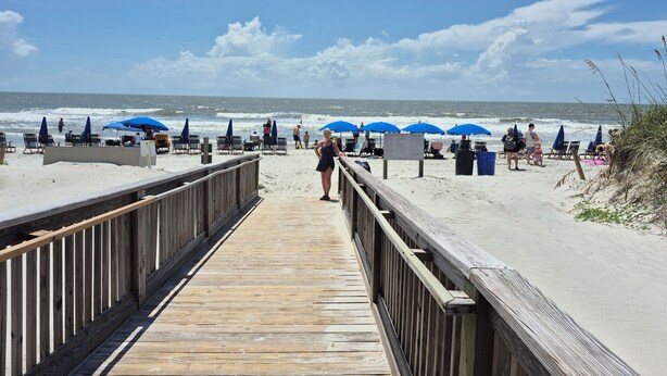 Une promenade en bois menant à une plage où se trouvent des parasols bleus et des chaises longues.