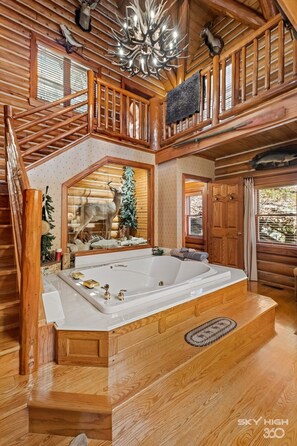 Interior espacioso de una cabaña de troncos con una amplia escalera de madera, un jacuzzi central y decoración rústica que incluye cuernos y trofeos de animales.