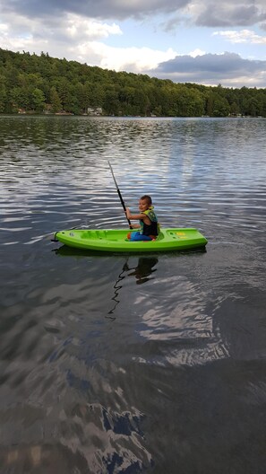 Ein Kind in einem grünen Kajak auf einem ruhigen See mit bewaldeter Uferlinie im Hintergrund.