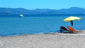 A beach with a boat in the water, a mountain range in the background, and a yellow umbrella on the sand.