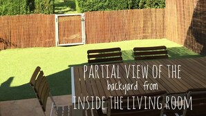 Vista parcial de un jardín trasero desde el interior de una sala de estar, con una terraza de madera, una cerca de bambú y un seto cuidado.