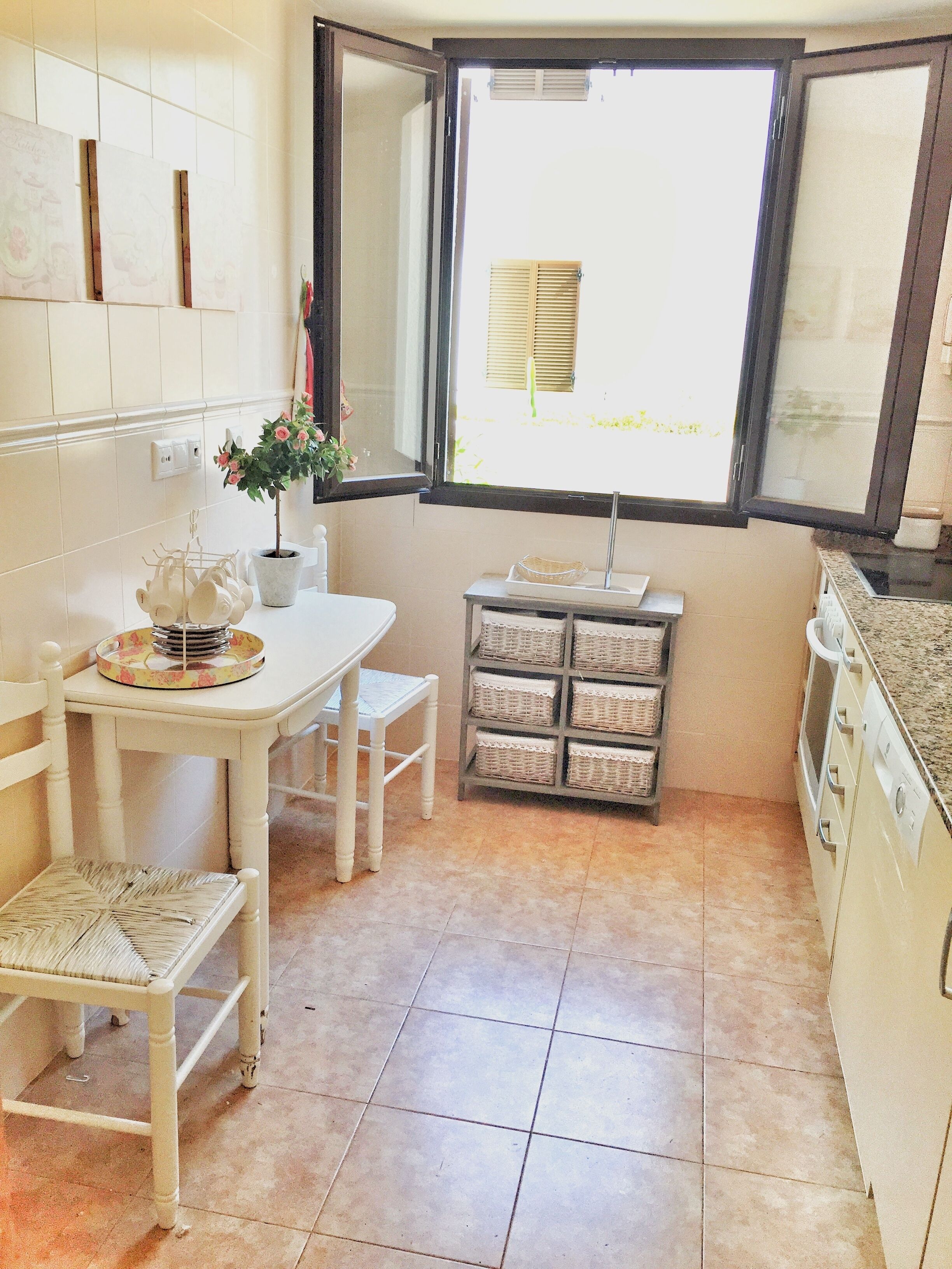Una cocina con una mesa de comedor, sillas, una planta en maceta, una ventana con persianas y un pequeño estante con cestas.