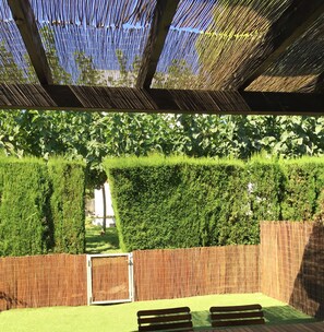 Vista parcial de un jardín trasero desde el interior de una sala de estar, con una terraza de madera, una cerca de bambú y un seto cuidado.