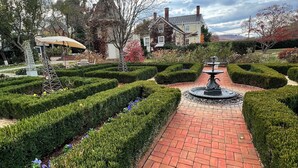 Un chemin pavé de briques mène à une fontaine, qui est entourée de haies soigneusement taillées et d’une variété de plantes.