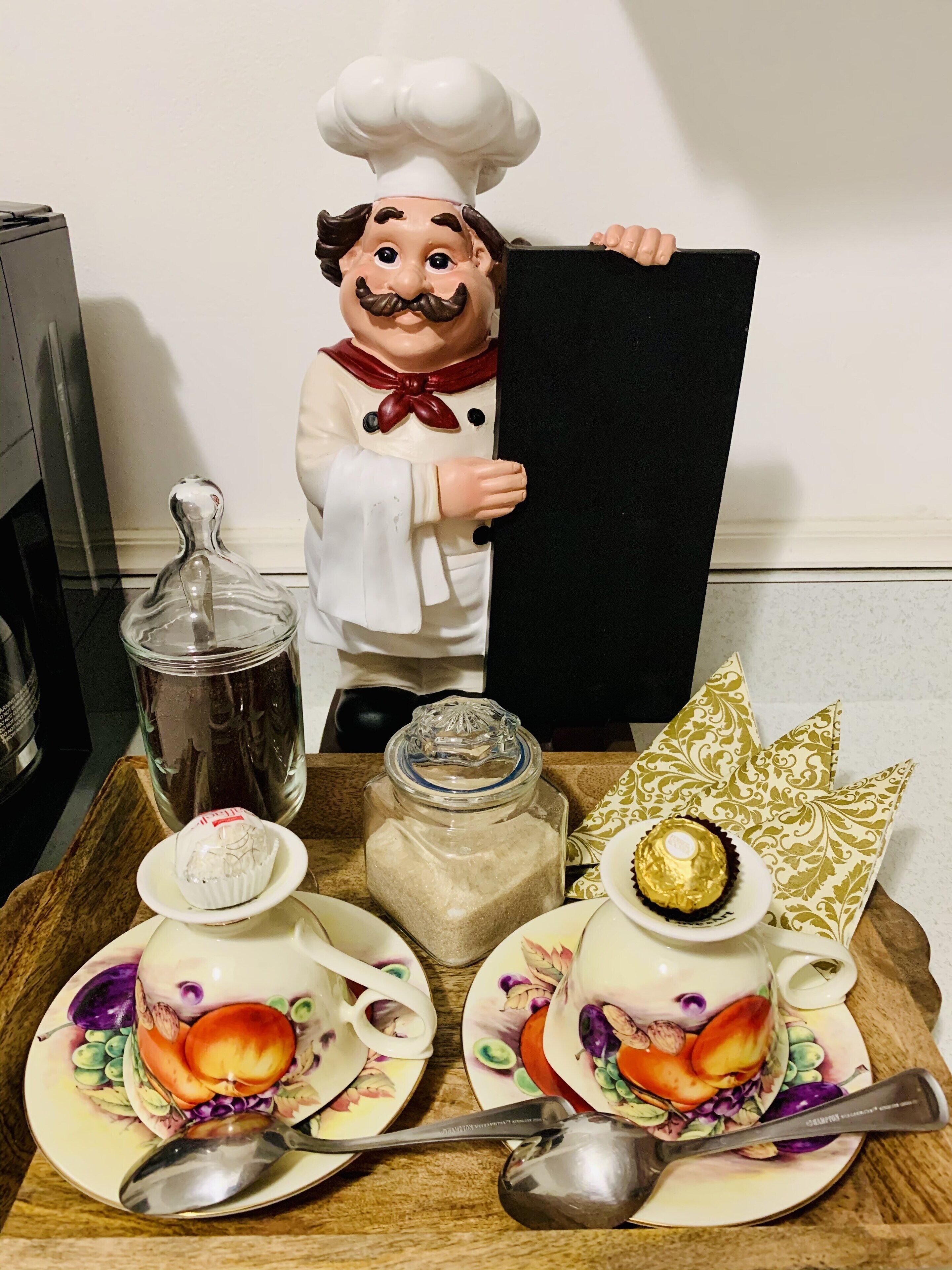 Un comptoir de cuisine agrémenté d’une statuette décorative représentant un chef, d’un bocal en verre, d’un récipient à sucre, de deux tasses à thé ornées de motifs floraux et d’un coussin décoratif.