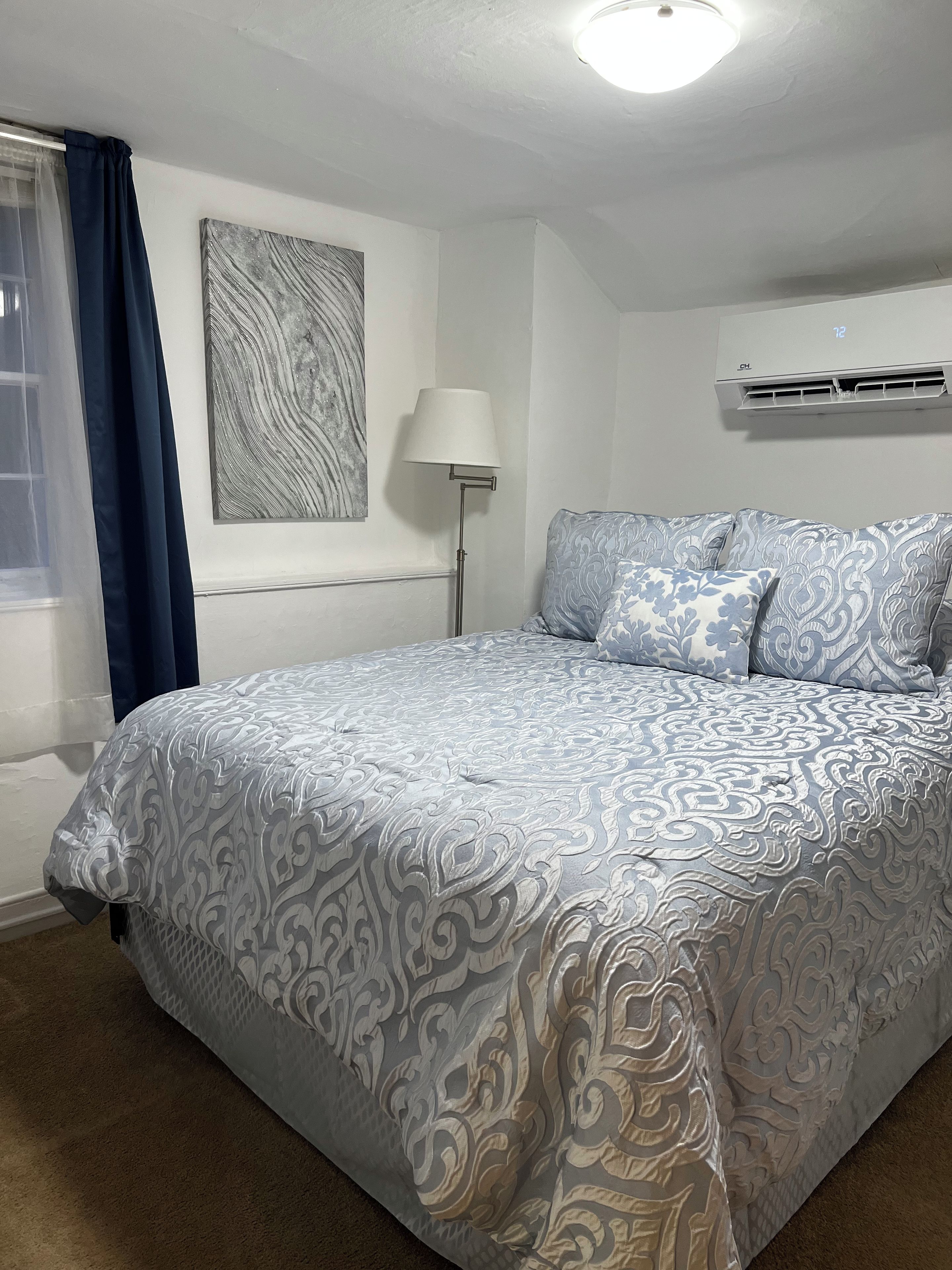 A neatly made bed with patterned bedding, a floor lamp, a wall-mounted air conditioner, and a window with curtains.