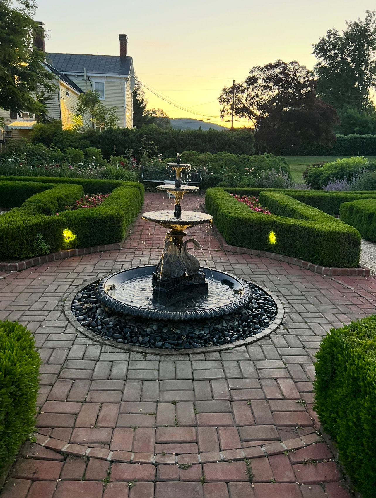 Un jardin avec une fontaine centrale, entouré de haies soigneusement taillées et d’un chemin en briques.