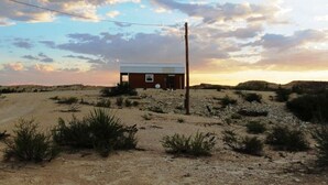 A small building with a flat roof and a single door, surrounded by sparse vegetation and a dirt road. - Sherri’s Terlingua Cabin on the Creek: Easy Access (Terlingua)