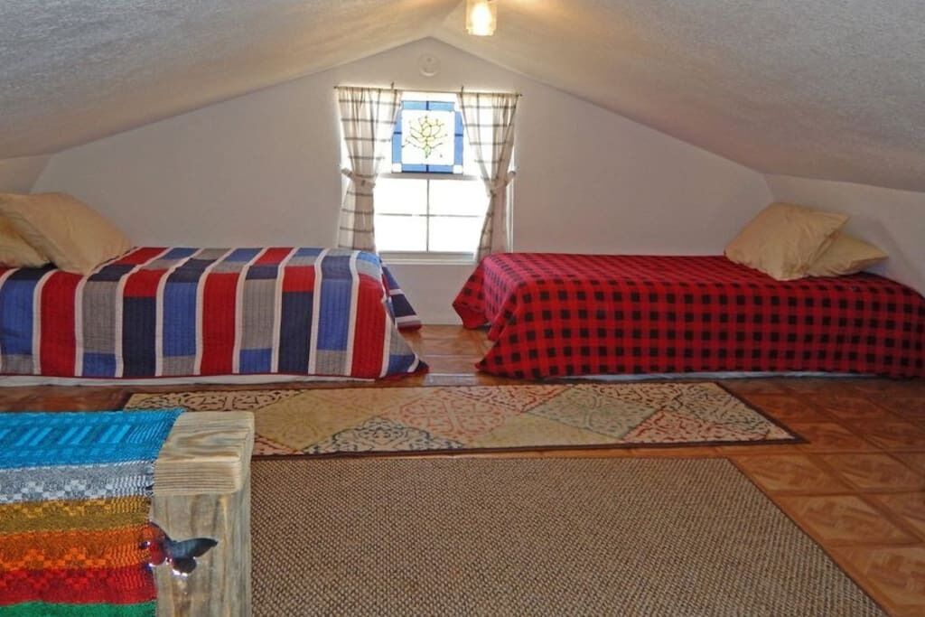A small attic bedroom with two beds, a window with curtains, and a variety of rugs on the floor.