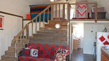 Un escalier menant à une mezzanine où se trouvent une horloge, un canapé rouge et gris à motifs et un coin cuisine.