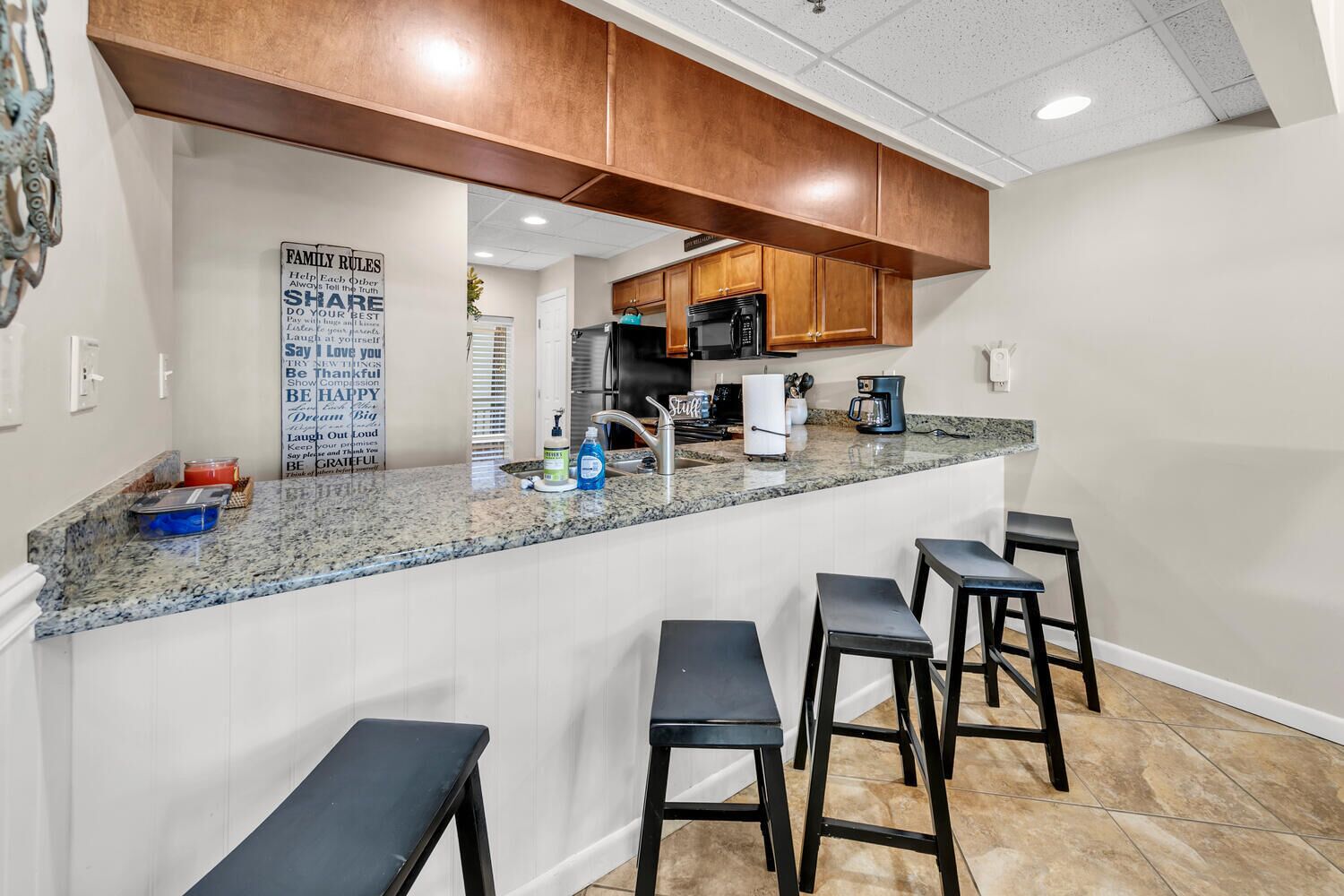 A kitchen with granite countertops, wooden cabinets, a stainless steel refrigerator, a coffee maker, and a bar stool area.
