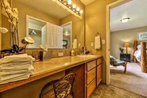 Un bagno con un mobile vanità, lavabo e specchio. Asciugamani sono impilati sul ripiano, una sedia e un letto si intravedono sullo sfondo.