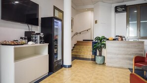 A hotel lobby with a reception desk, a refrigerator, a flat-screen TV, a staircase, and a potted plant. - Negresco Gran Vía (Madrid)