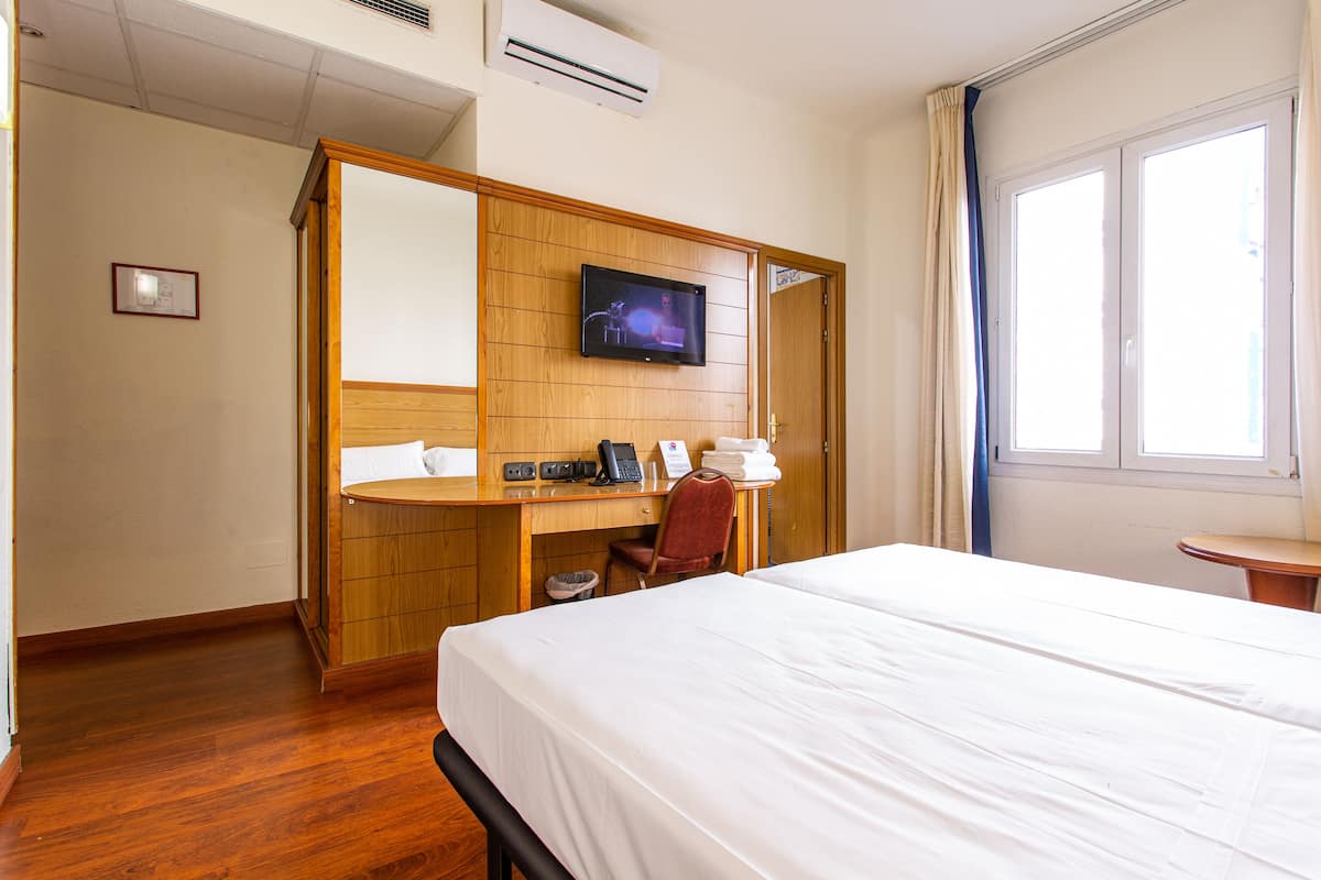 A hotel room with a bed, a desk with a computer, a TV, and a window.