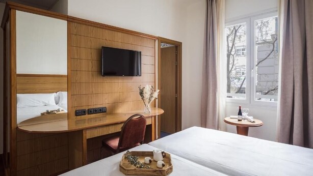 A hotel room with a wooden desk, a flat-screen TV, a bed, and a small table with a bottle and glasses. - Negresco Gran Vía (Madrid)