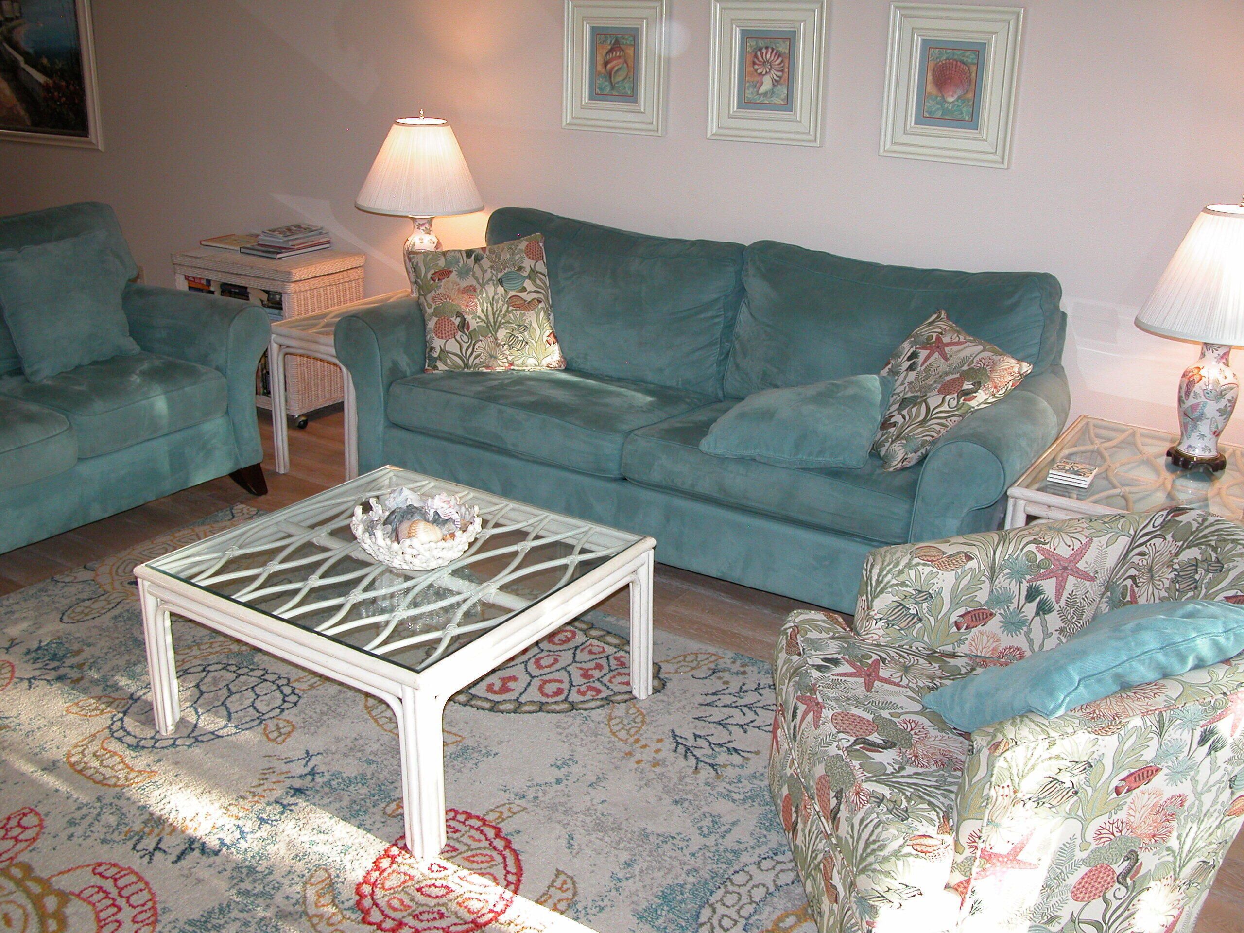 A living room with a teal sofa, patterned cushions, a glass coffee table, and a floral-patterned chair.