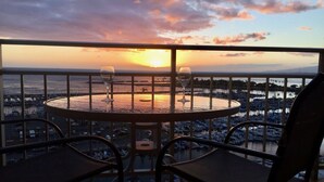 A balcony with a round table and two chairs, overlooking a marina with boats and a sunset. - Deluxe Waikiki Ocean Front Suite - Ilikai (Honolulu)