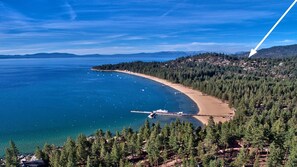 An aerial view of a coastal area with a beach, dense forest, and a pier.