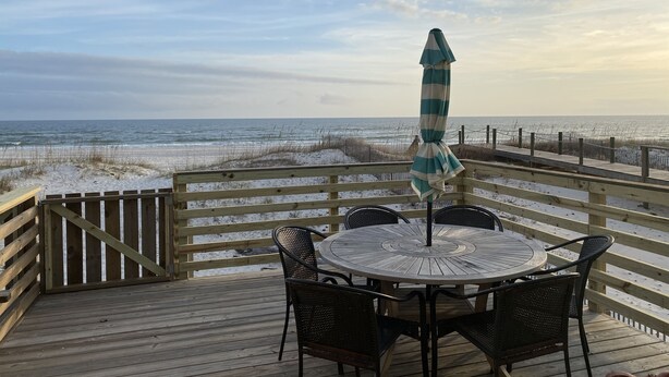A wooden deck with a round table and chairs, overlooking a beach with a fence and an umbrella. - 3BR Duplex ON The Beach, Sleeps 11 Comfortably (Gulf Shores)