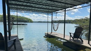 A wooden dock extending into a calm lake, with a hammock and chairs under a metal roof.
