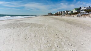 A wide sandy beach with footprints, a clear blue sky, and a row of houses with green roofs and white exteriors along the shoreline. - So Close to the Beach & Pool! (Pawleys Island)