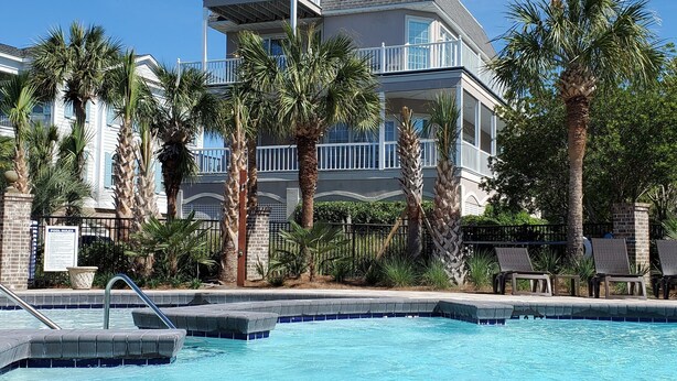 A pool area with a clear blue pool, surrounded by palm trees and a multi-story building with balconies. - So Close to the Beach & Pool! (Pawleys Island)