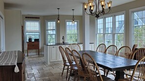 A spacious dining area with a large wooden table and chairs, a chandelier, and large windows offering views of the outside. - So Close to the Beach & Pool! (Pawleys Island)