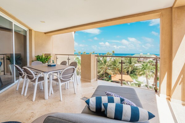 Ein Balkon mit Esstisch und Stühlen, eine Sitzecke mit gestreiften Kissen und Blick auf das Meer und Palmen.