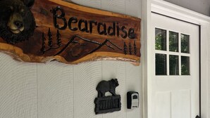 Un letrero de madera que dice 'Almost Paradise' y presenta una talla de oso, un cartel de bienvenida y una puerta con un ventanal.