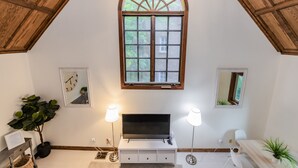 A room with a sloped ceiling, a large arched window, a flat-screen TV on a white console, two floor lamps, and a potted plant. - Castle like home in SW PDX, close to downtown! (Portland)