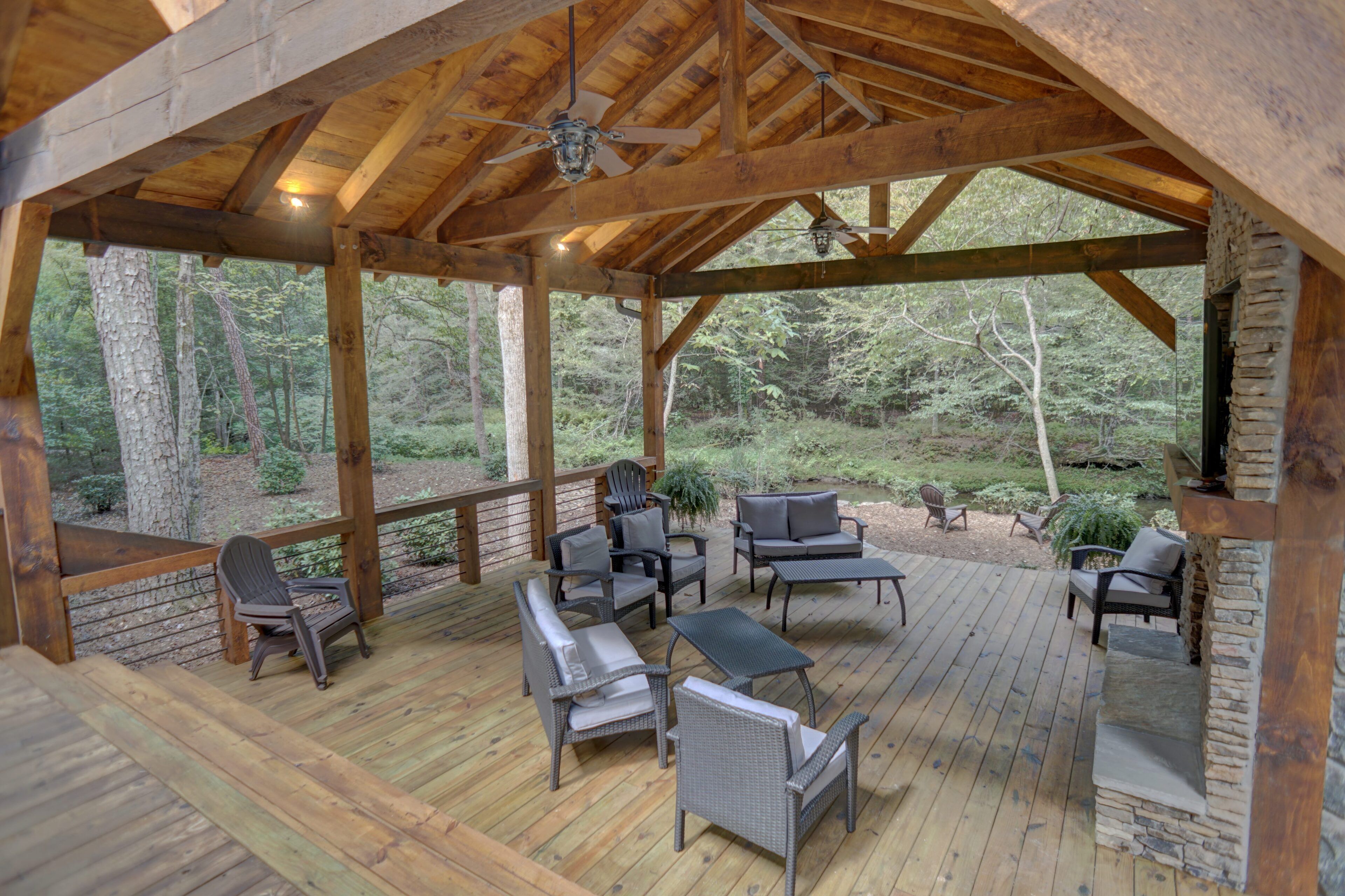 Une terrasse extérieure couverte, dotée de poutres en bois et d’un toit de chaume, aménagée avec des fauteuils et une table en osier, et entourée d’arbres.
