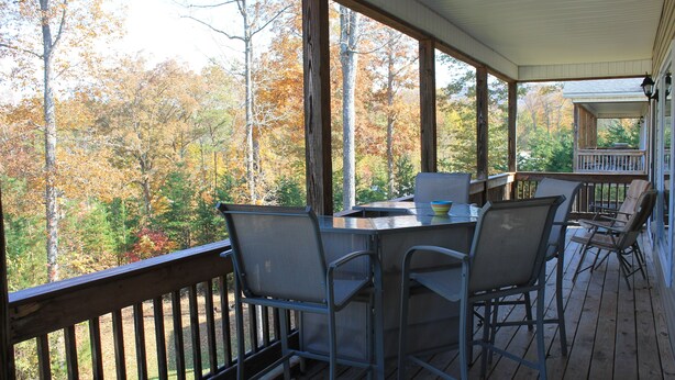 Um terraço coberto ao ar livre com mesa e cadeiras, com vista para uma área arborizada.