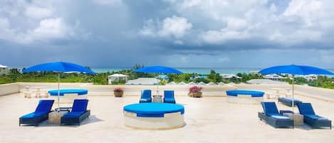 A rooftop pool area with blue lounge chairs and umbrellas, a clear view of the ocean, and a cloudy sky.