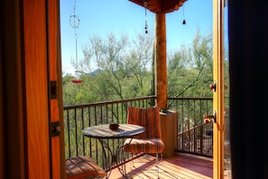 A wooden deck with a round table and two striped chairs, a wooden railing, and a view of trees and mountains in the background.