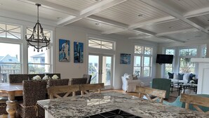 A spacious kitchen with a large island, wooden chairs, and a dining table. There are large windows and artwork on the walls.