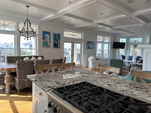 A spacious kitchen with a large island, wooden chairs, and a dining table. There are large windows and artwork on the walls.