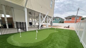 Un campo da golf in erba sintetica ben curato, con tre bandiere, circondato da una recinzione bianca e adiacente a un edificio con una veranda coperta.